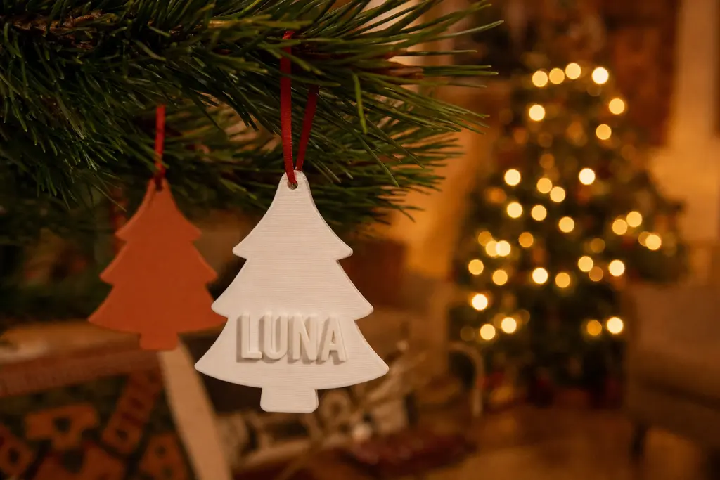 Adorno navideño impreso en 3D con forma de pino, en blanco hueso mate y con el apellido LUNA en relieve, colgado de una rama con listón rojo junto a otro adorno terracota, con las luces cálidas del árbol al fondo.